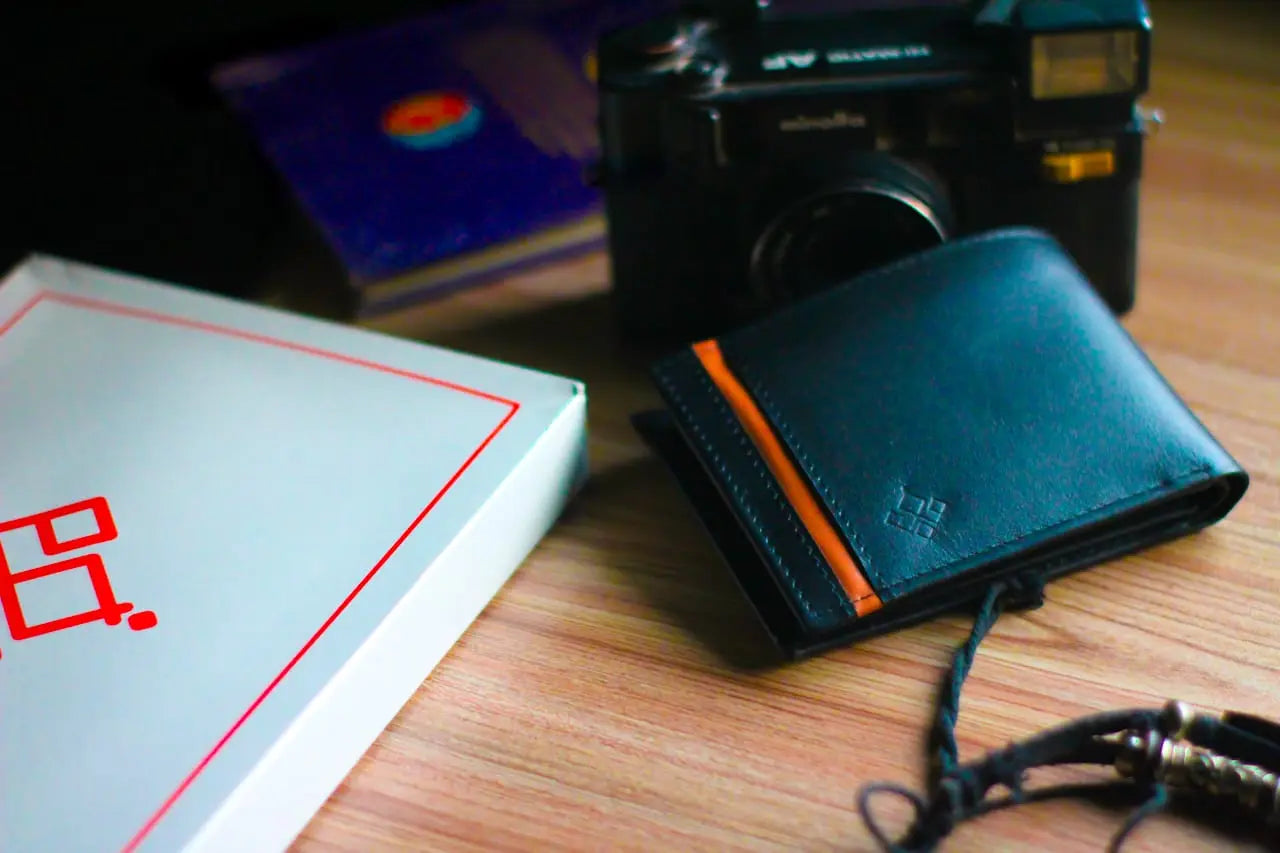 Black leather wallet with orange accent on a wooden surface next to a camera and book.